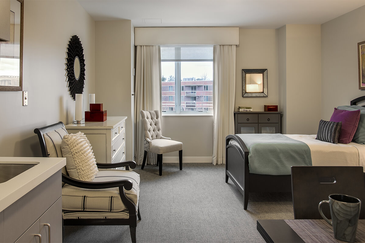 Interior view of a resident bedroom at an independent living facility. The bedroom features light colored walls with a light grey carpet floor. Dark walnut bedroom furniture is seen, with several small dressers, upholstered chairs, and a bed.