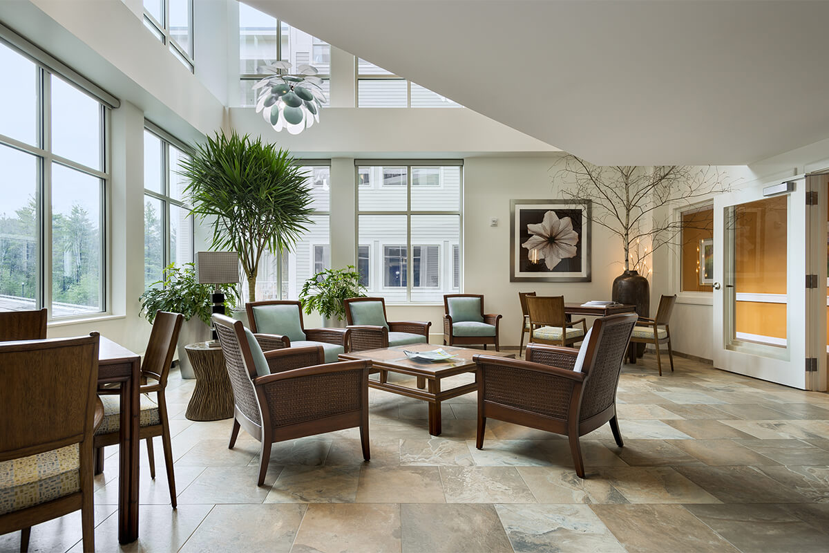 Interior photo of a sunroom area at an independent living facility. The room is bright and airy with neutral colored stone tile floor and light colored walls. Wood and upholstered chairs are gathered around a low rectangular coffee table.
