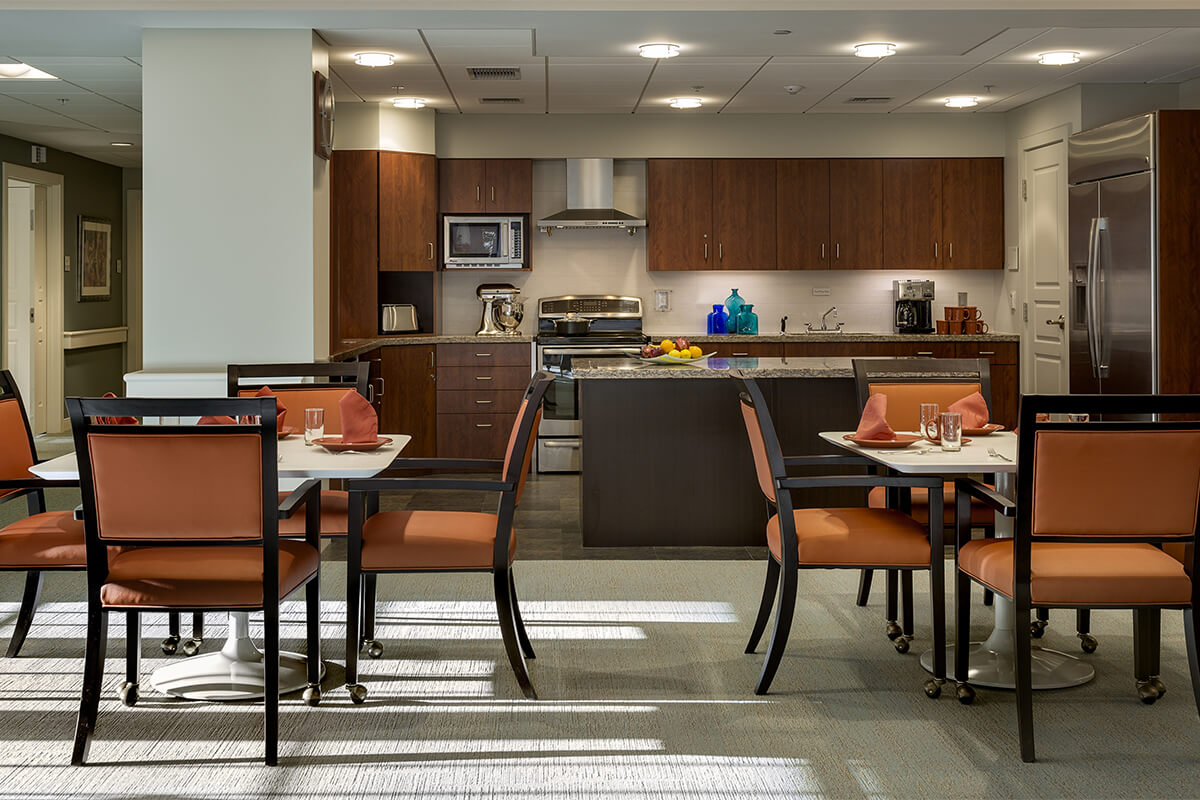 Interior photo of a dining area at an enhanced independent living facility. Two rectangular tables are seen in the foreground with orange upholstered dining chairs around them. In the background, a kitchen area with walnut cabinets and kitchen island.