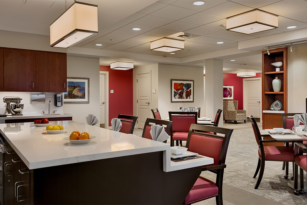 Interior view of an assisted living dining area. A kitchen island in the foreground shows dark wood and red upholstered chairs around it.
