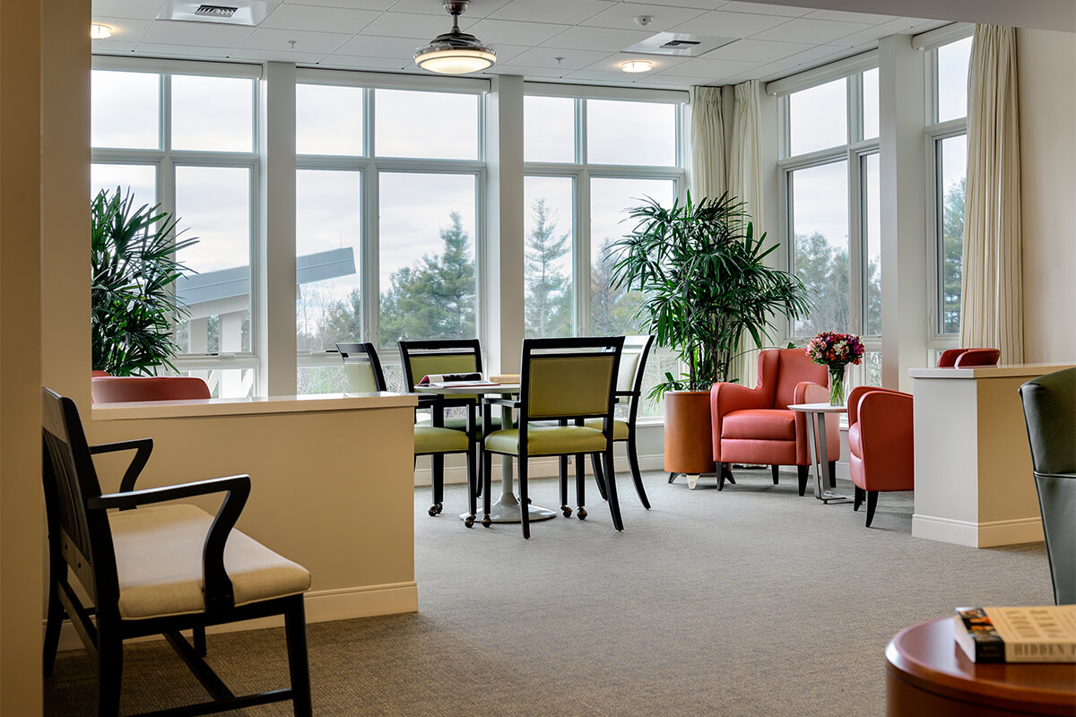 Interior view of a memory care household area at a senior living facility. Green and walnut dining chairs are around a small table, and red leather chats are in a seating area by large glass windows.