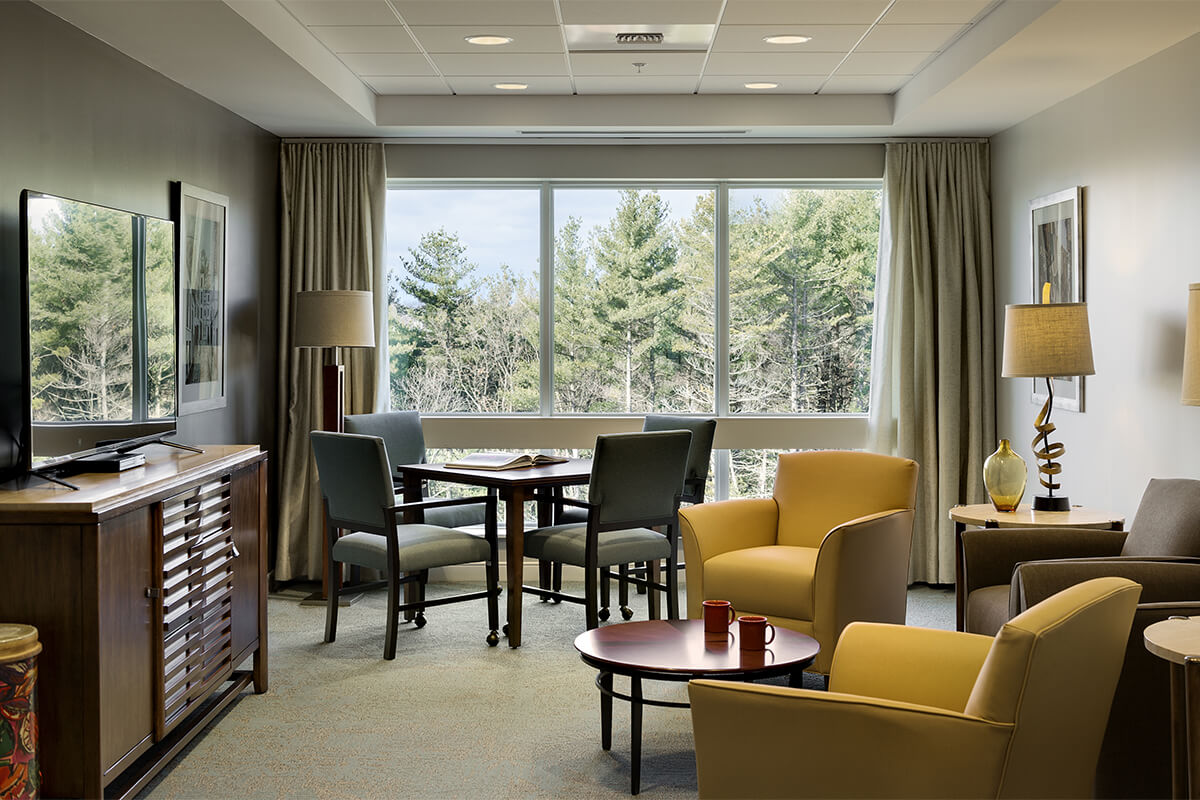 Interior view of a living den in a resident's living unit at a senior living facility. The photo shows a back wall with large windows showing a wooded area. Yellow and light blue upholstered chairs, small tables, and a TV on a stand are seen within the room.