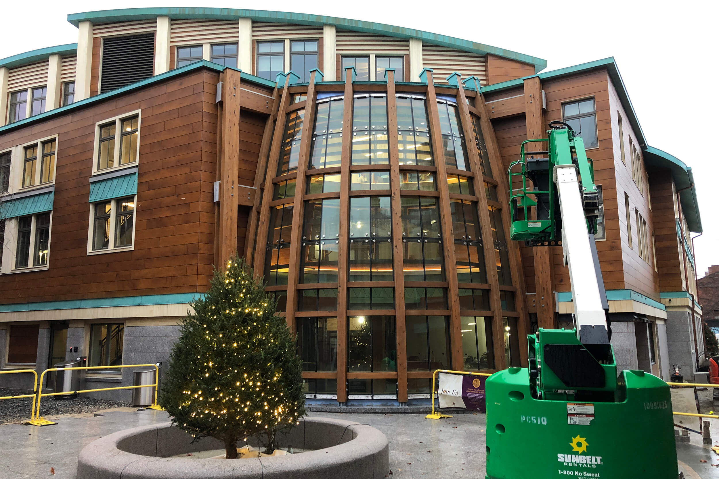 Exterior daytime photo of the facade of a mixed-material (glass, wood, brick) building. In front of the building is a Christmas tree and a crane.