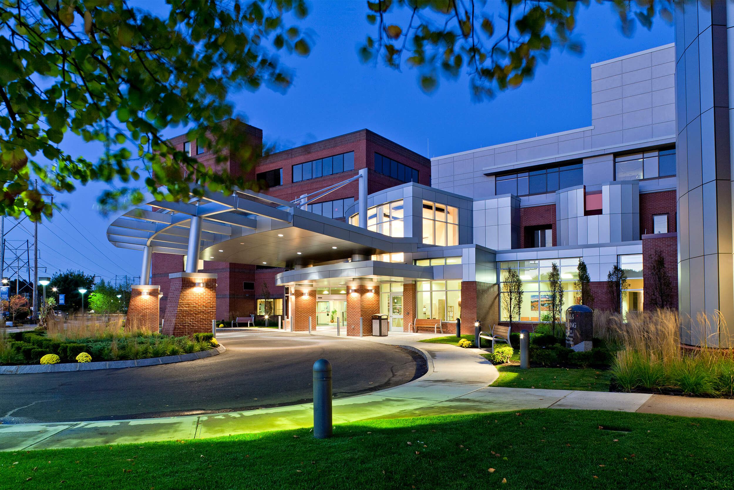 Exterior view of a hospital entrance. The photo is taken at dusk with low lighting - lights on the exterior and interior of the building illuminate it. The building features a mix of concrete and redbrick facade treatments, and a driveway curves around the entrance area.