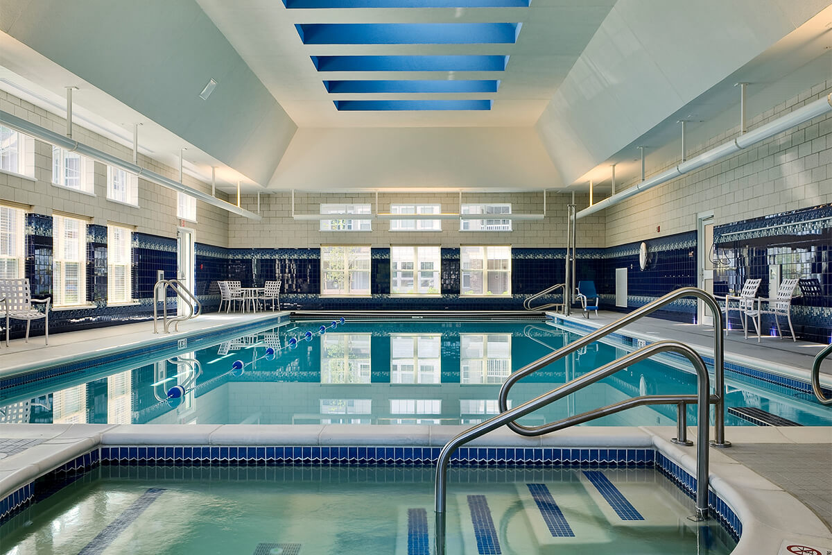 Interior photo of an indoor pool at an assisted living facility. The rectangular pool has clear blue water and there are a few white pool chairs along the sides.