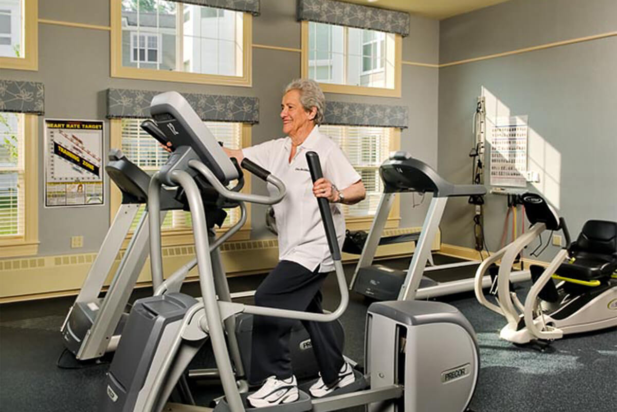 A senior woman enjoys working out on an elliptical machine at the fitness center of a senior living facility.