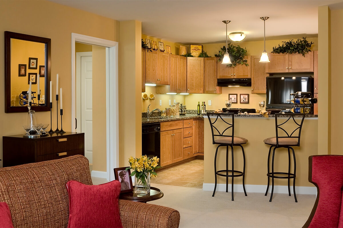 Interior photo of a living space at an independent senior living facility. The view is from a living room area into a small kitchen area, with neutral colored walls and a bistro countertop.