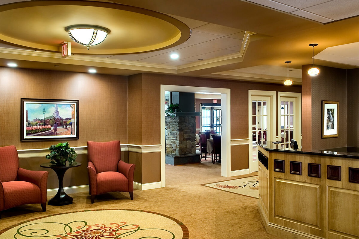 Interior view of a lobby at an assisted living facility. The room features neutral and light brown beige tones, with red upholstered seating. A reception desk can be seen on the right side, and a look into the room beyond shows a stone fireplace.