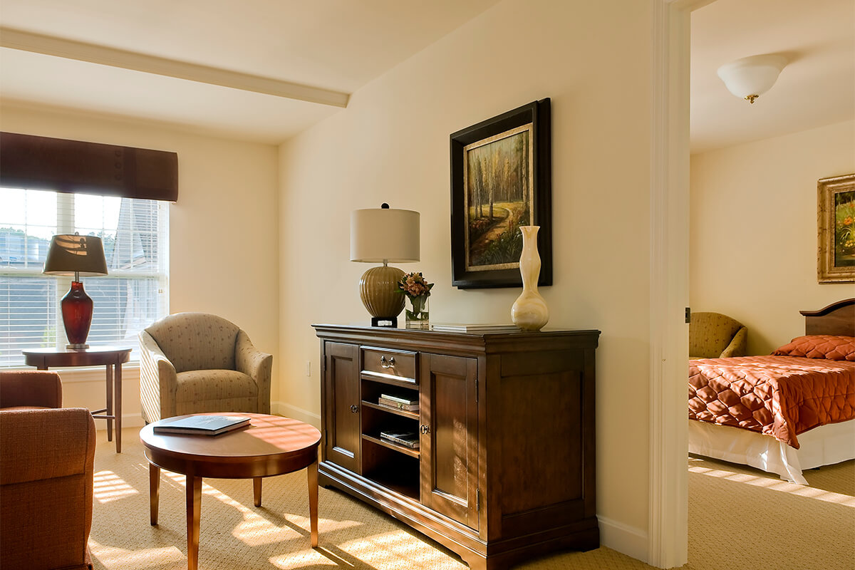 Interior photo of an apartment space at a senior living facility. The room is bright and cozy with natural light coming from a window on the far wall. The walls and flooring are neutral colored, with a dark wood console cabinet against one wall. An upholstered chair and small end table is seen, and the camera reveals a little bit of the bedroom in an adjacent room.