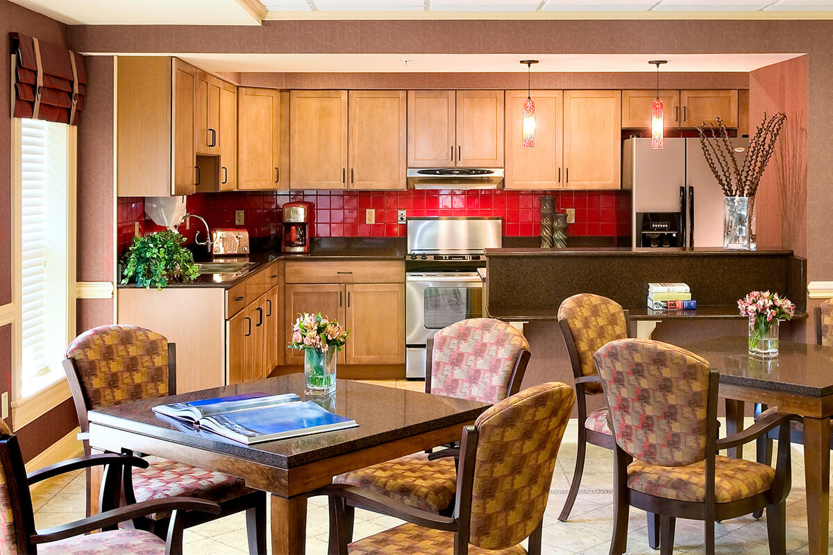Interior photo of a kitchen dining area at a senior living facility. Wood tables and patterned upholstered dining chairs are seen in the foreground, and a small kitchen area with red tile backsplash is in the background with light wood cabinets.