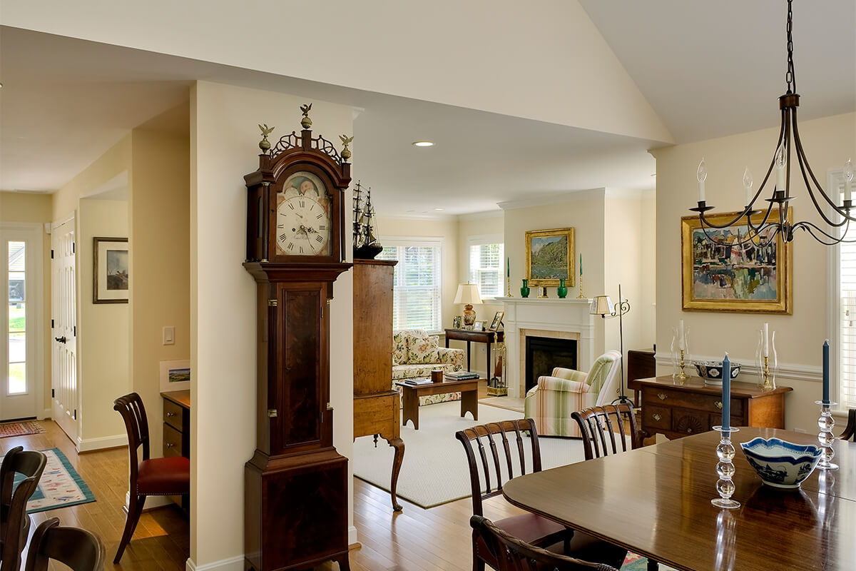 Interior view of a living room in a cottage unit at a senior living facility. The walls and flooring are neutral light colors, with dark wood furniture accenting the space. A grandfather clock, dining table, and living room furniture set are seen.