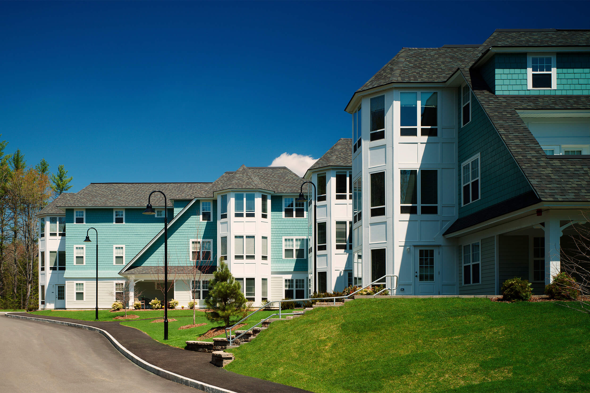Exterior daytime photo of a senior living facility. The multi-unit buildings feature light blue siding with white trim accents, and grassy lawns.