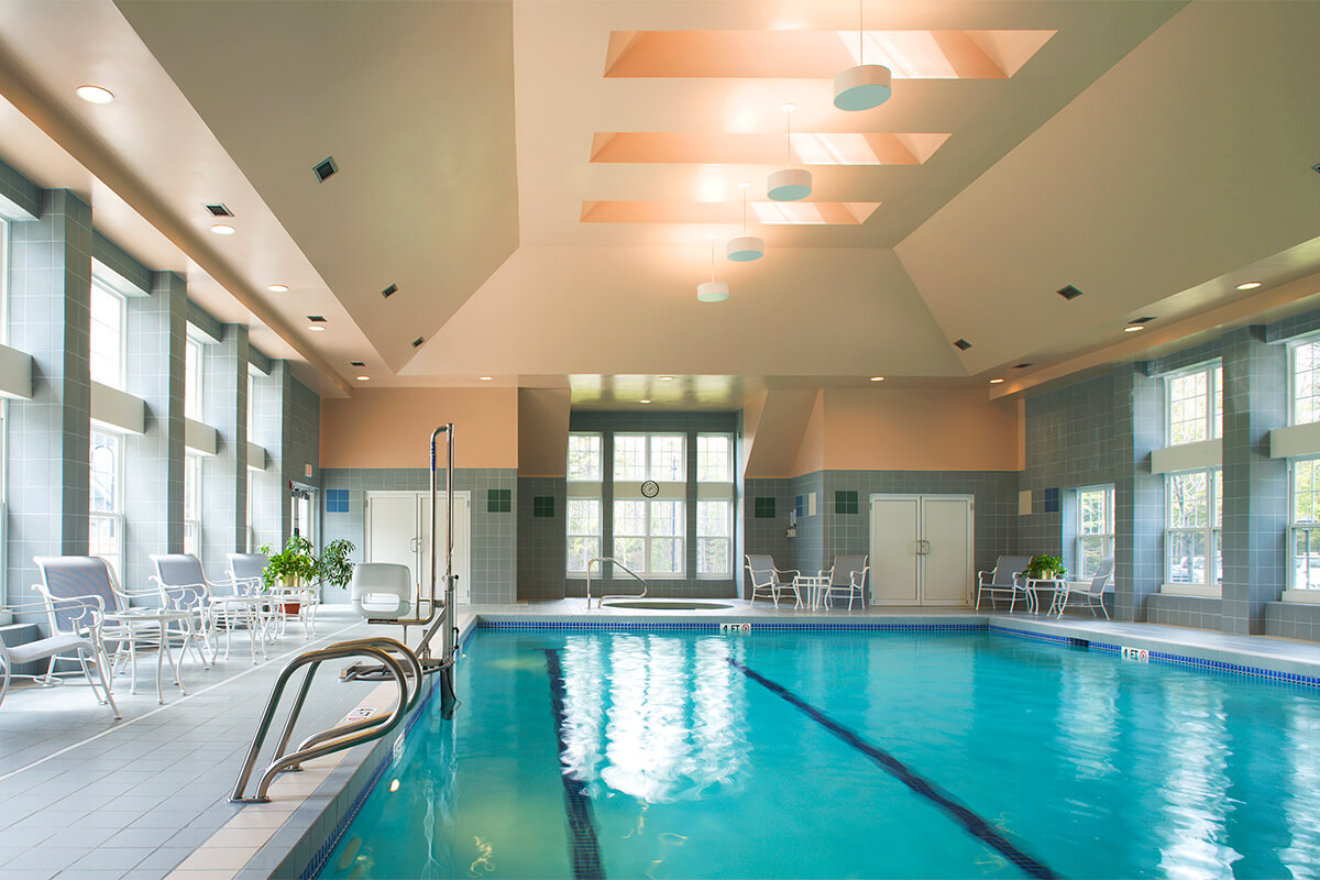 Interior photo of an indoor pool area at a senior living facility. The rectangular pool features clear blue water, and the room has lots of natural light from large windows and white metal chairs on either side of the pool.