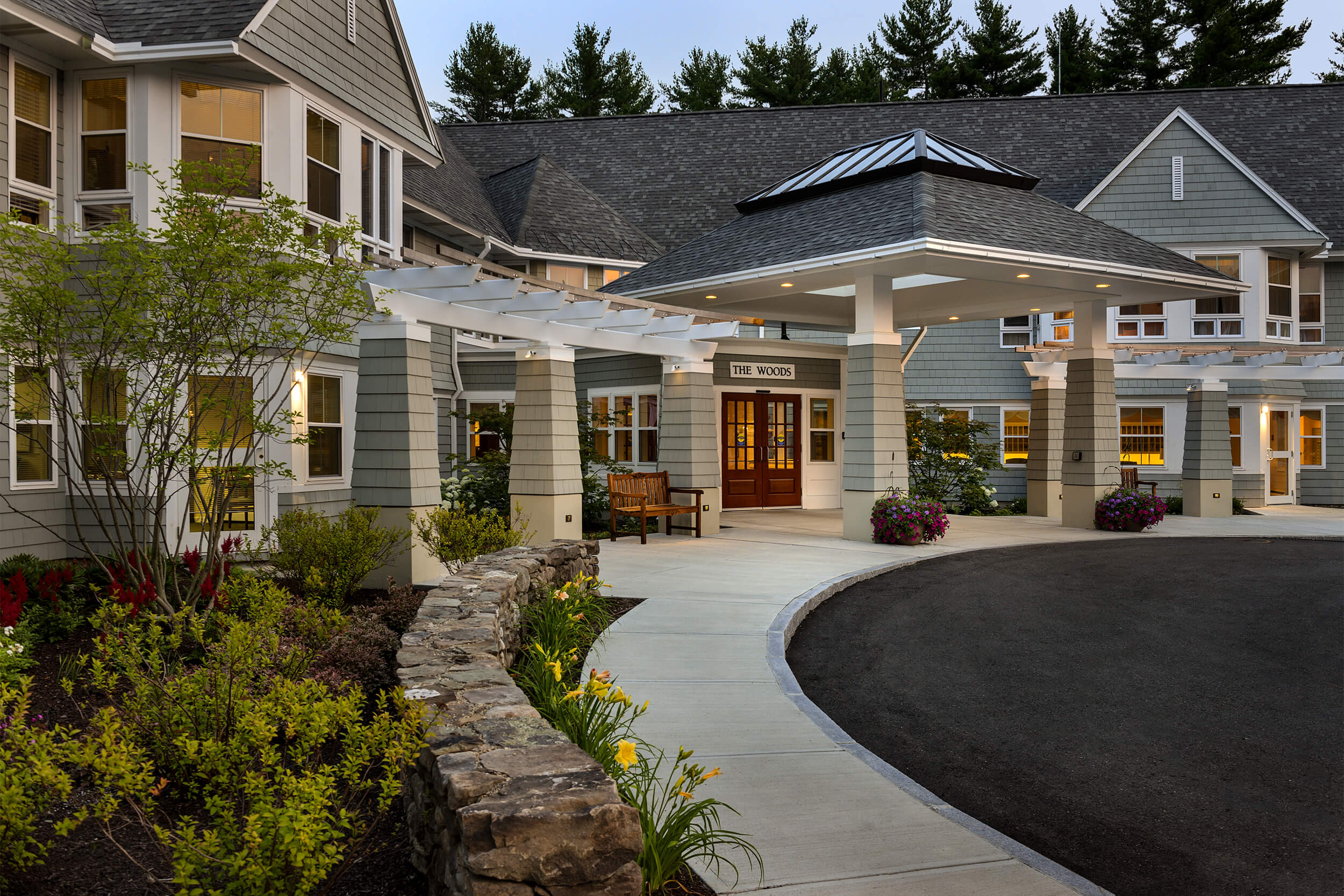 Exterior daytime photo of the entrance of a senior living facility. The building has grey siding and white trim, with plantings and mulch beds adjacent to the facade of the building and a curved driveway.
