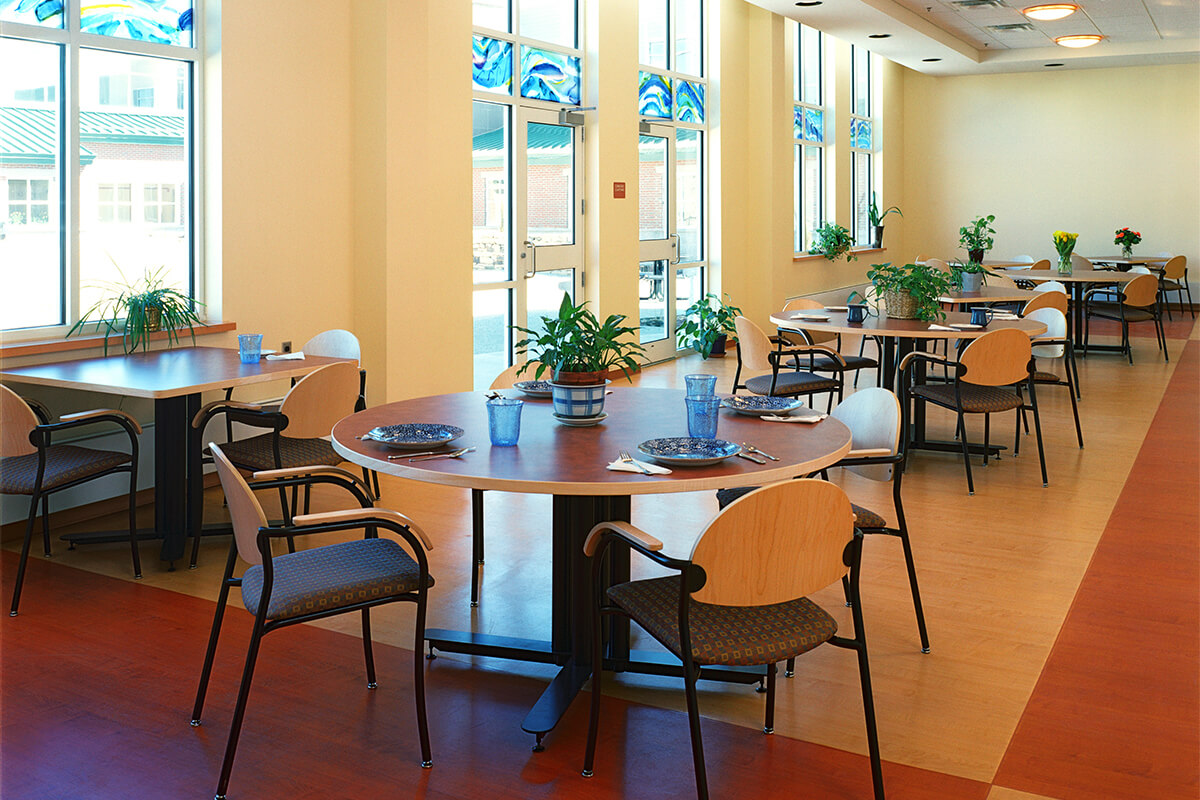 Interior view of a dining cafeteria at a psychiatric center. The room has neutral colored walls with a dark and light wood floor, and large windows and doors letting in natural light. Several round dining tables with place settings and dining chairs can be seen. Potted plants on the tables and windowsills add natural greenery to the space.