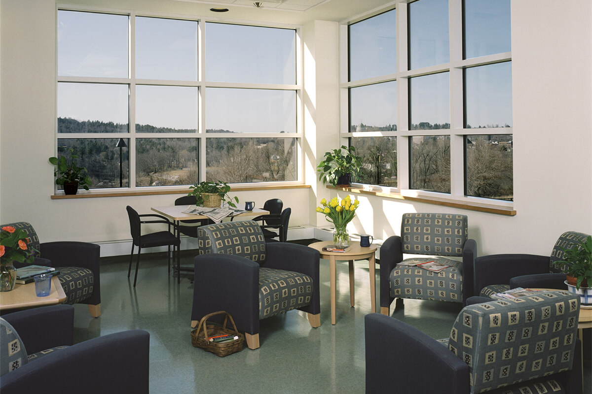 Interior photo of a sitting area in a day room at a psychiatric care center. The photo shows the corner of a room where rows of large windows meet in a corner, and several comfortable blue and green patterned chairs gathered around. There are small tables, some craft and reading supplies, and various small potted plants throughout the area.