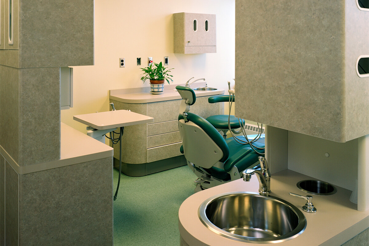 Interior view of a dentist office as part of a psychiatric care center. The office features neutral colored walls and cabinets, and a green dentists chair and sink are seen.
