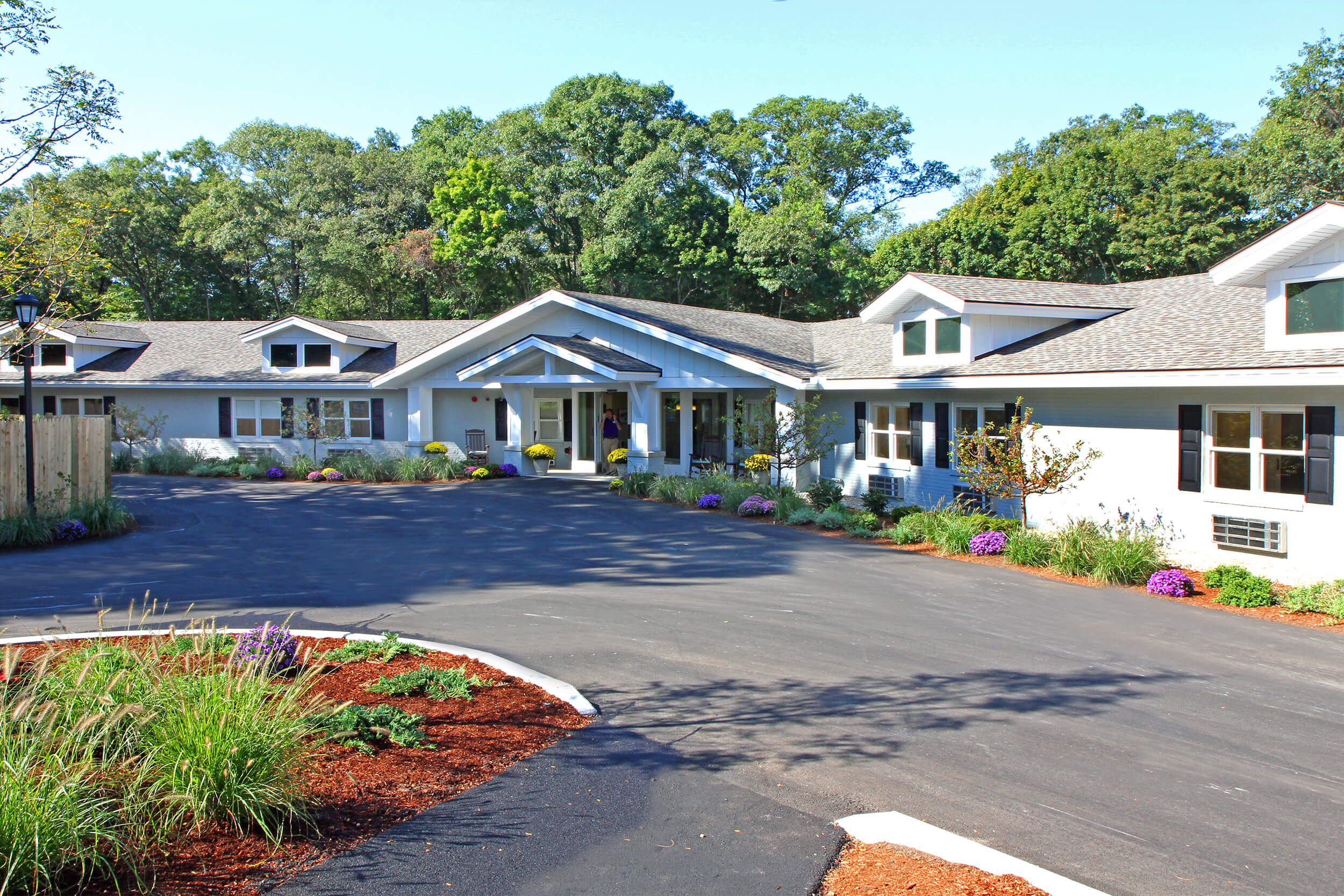 Exterior daytime view of entrance to a senior living facility. THe building is low and long, featuring white siding with black window shutters. Low bushes and plants are in the mulch along the front edge of the building, and a circular driveway area is seen in the front.