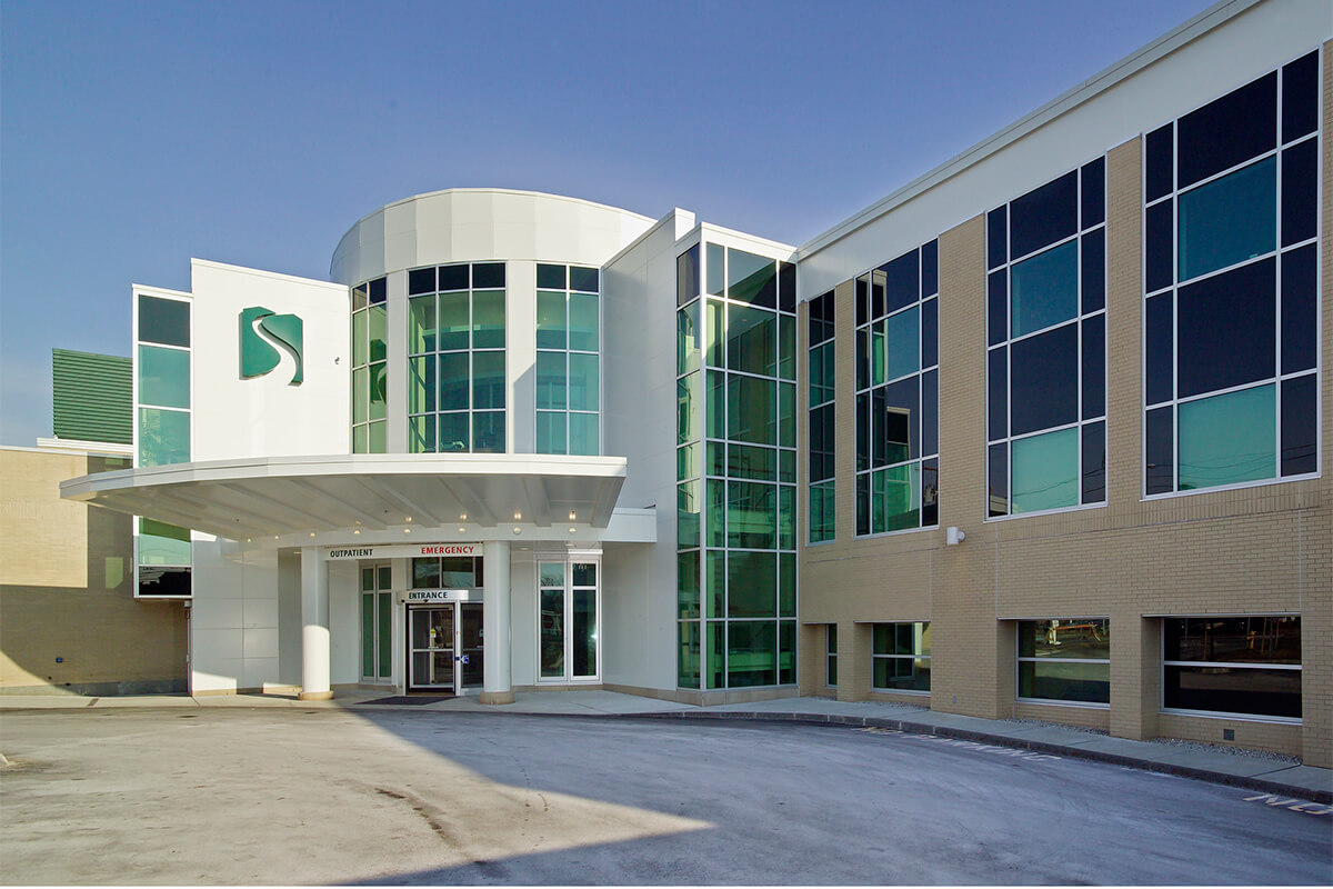 Exterior daytime photo of the entrance of an emergency department at a medical center. The white multi-floor building features green tinted large windows.
