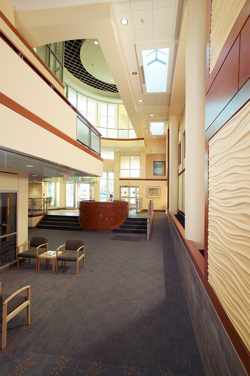 Interior view of the atrium lobby at a medical center. The floor is a dark grey carpet with several waiting area chairs seen in the photo, and the walls are a neutral beige with wood accents. The accent wall on the right features a sculptural wave pattern.