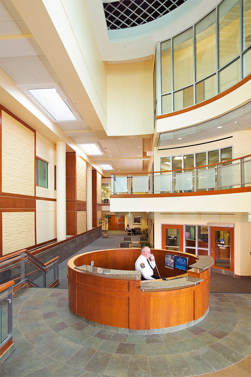 Interior photo of a main lobby area at a medical center. The photo shows a circular wooden security desk manned by a gentleman in a security officer uniform who is answering a phone. The floor features greyish-green tiles in a circular pattern, and the walls are a neutral beige with wood trim.