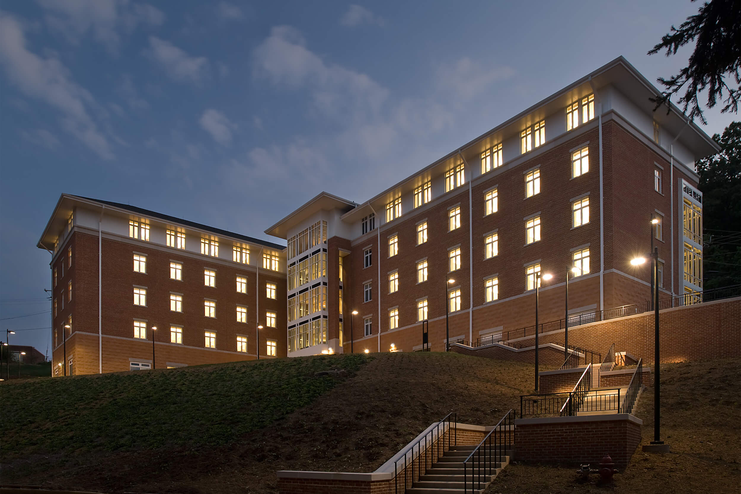 Exterior view of Kellogg House student residence building at University of Virginia. The photo is taken in the early evening and shows an angled view of the residence building and the stairway leading up to it. The building features a brown brick facade with white trim and windows, and lights are illuminating the building from within.