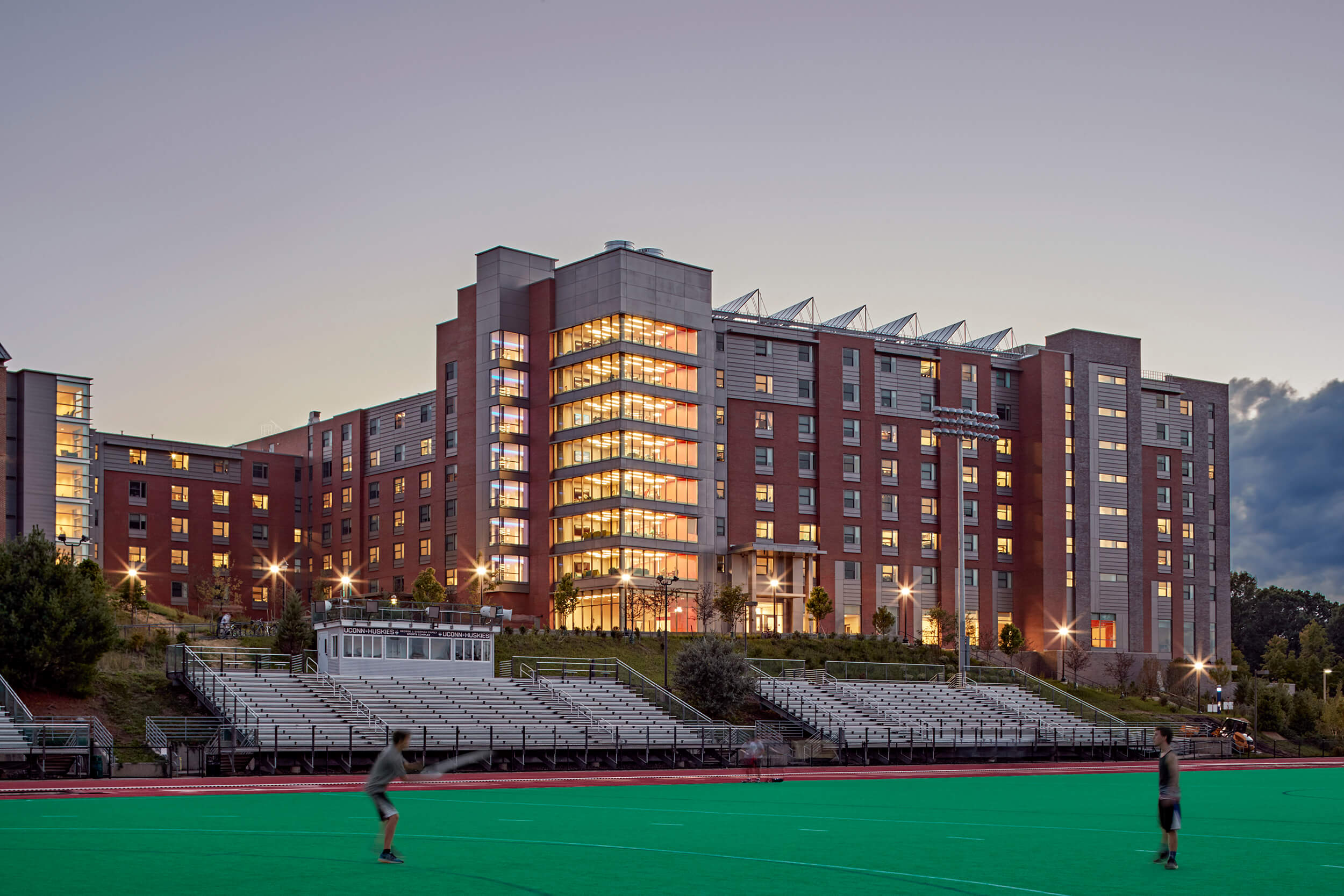 Exterior view of a college campus building as seen across an athletic field. The photo appears to be taken at dusk, with lighting illuminating the building from within.