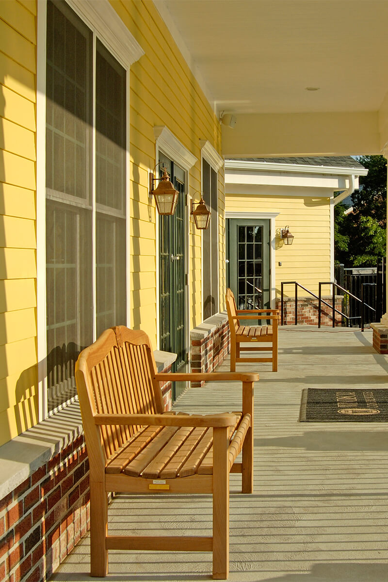 An exterior photo of a porch at an apartment complex clubhouse. The deck of the porch is a light grey with light brown benches on it.
