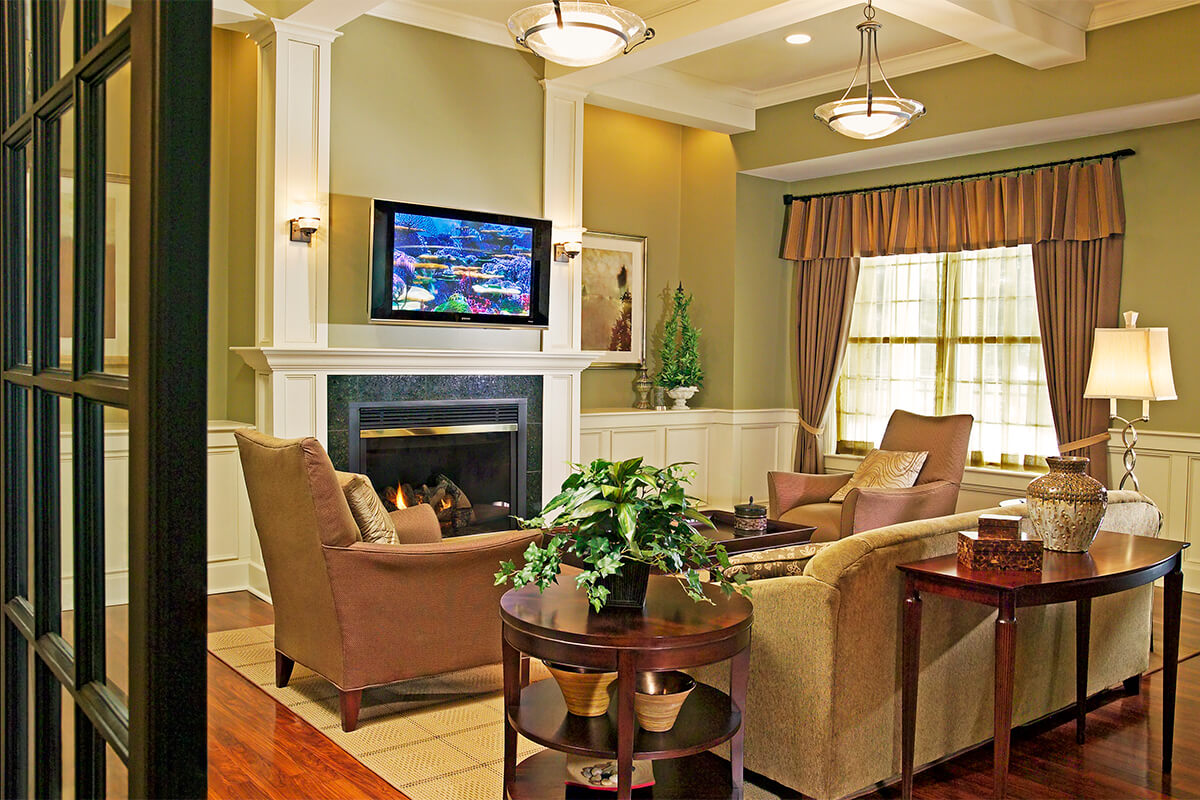 Photo of the interior of a clubhouse at an apartment complex. The photo shows a living-room like environment with two plush chairs and a couch gathered around a fireplace with a TV unit mounted above. The walls are a light green with white trim and wainscoting, light brown window curtains, and dark wood end tables and coffee table.