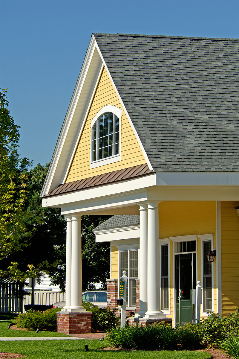 Detailed closeup view of the exterior of the entrance to a clubhouse at an apartment complex. The building has yellow siding with brick and white trim, and a flat walkway leading up to the entrance with small planted bushes on either side.