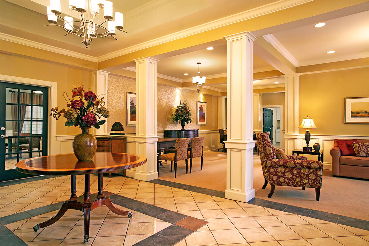 Photo of the interior of a lobby space at an apartment complex clubhouse. Light colored tiled floor meets a carpeted area, and the room is separated with white columns. A large dark wood circular table is in the center of the titled area with a vase of flowers. Various living room furniture (chairs, couches, end tables) are seen further back in the distance on the carpeted area.