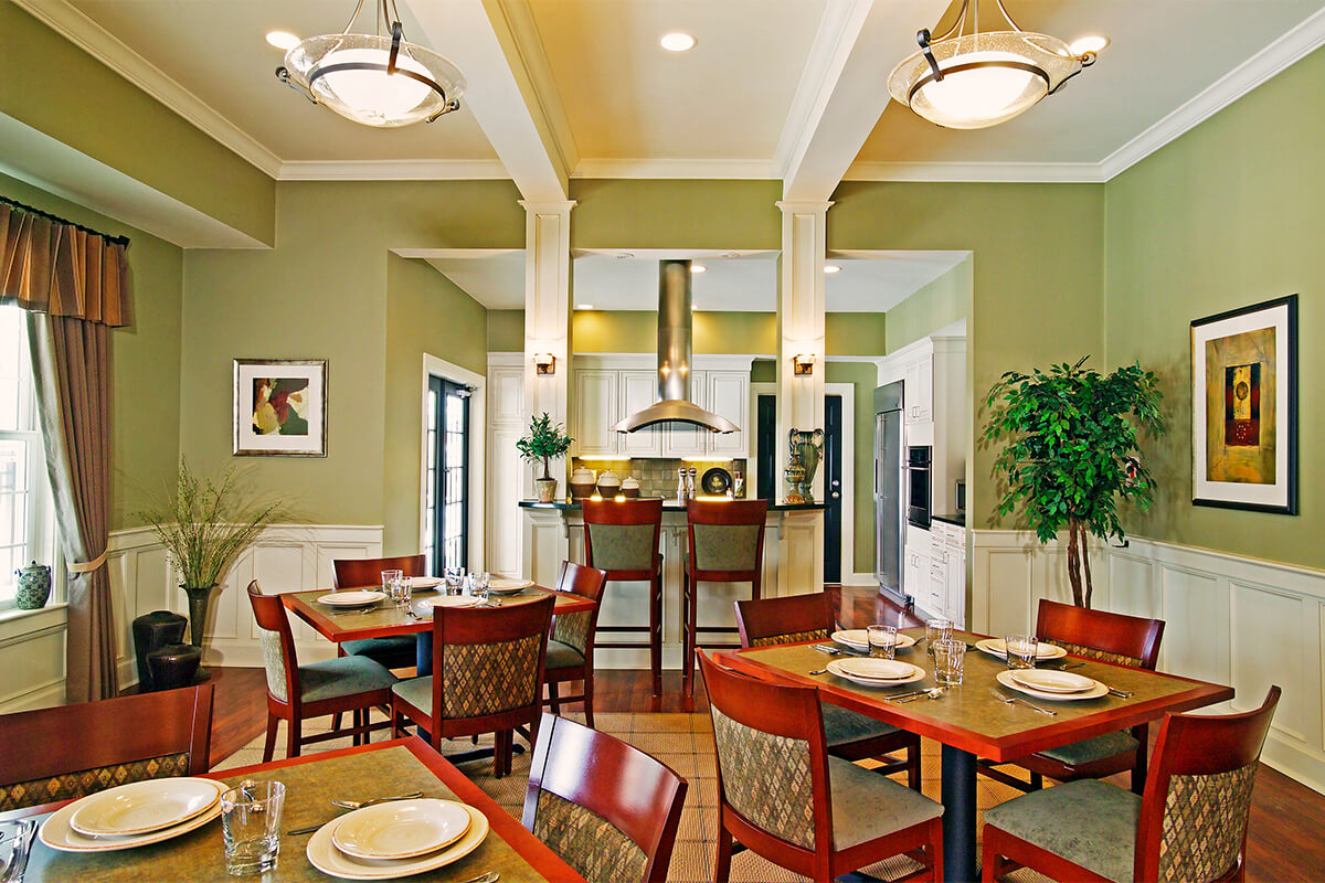 Photo of the interior of a community room at an apartment complex. The walls are a light green with white wainscoting and trim, and several square dining tables are seen in the foreground with chairs around them.