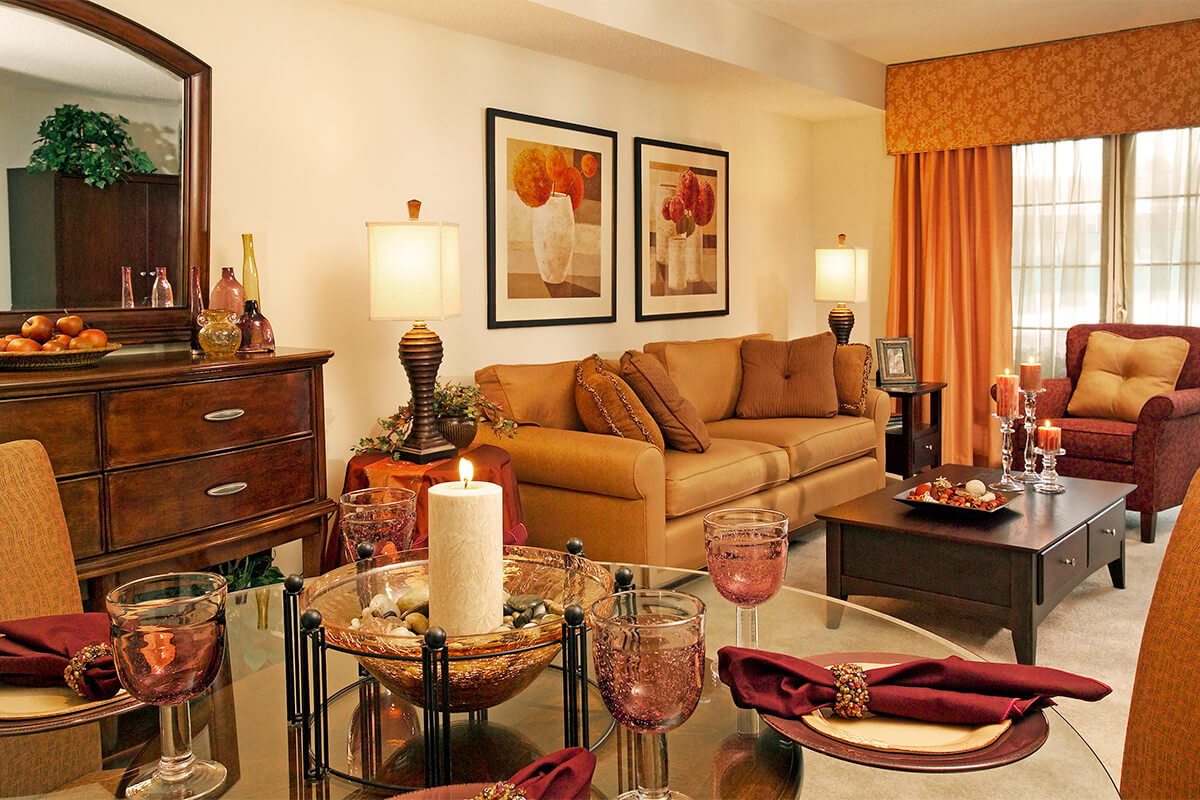 Photo of the interior of a model apartment unit living room. The living room features neutral walls, dark wood furniture, light orange and red couch and sitting chair, orange curtains, and a glass dining table with a candle centerpiece in the foreground.