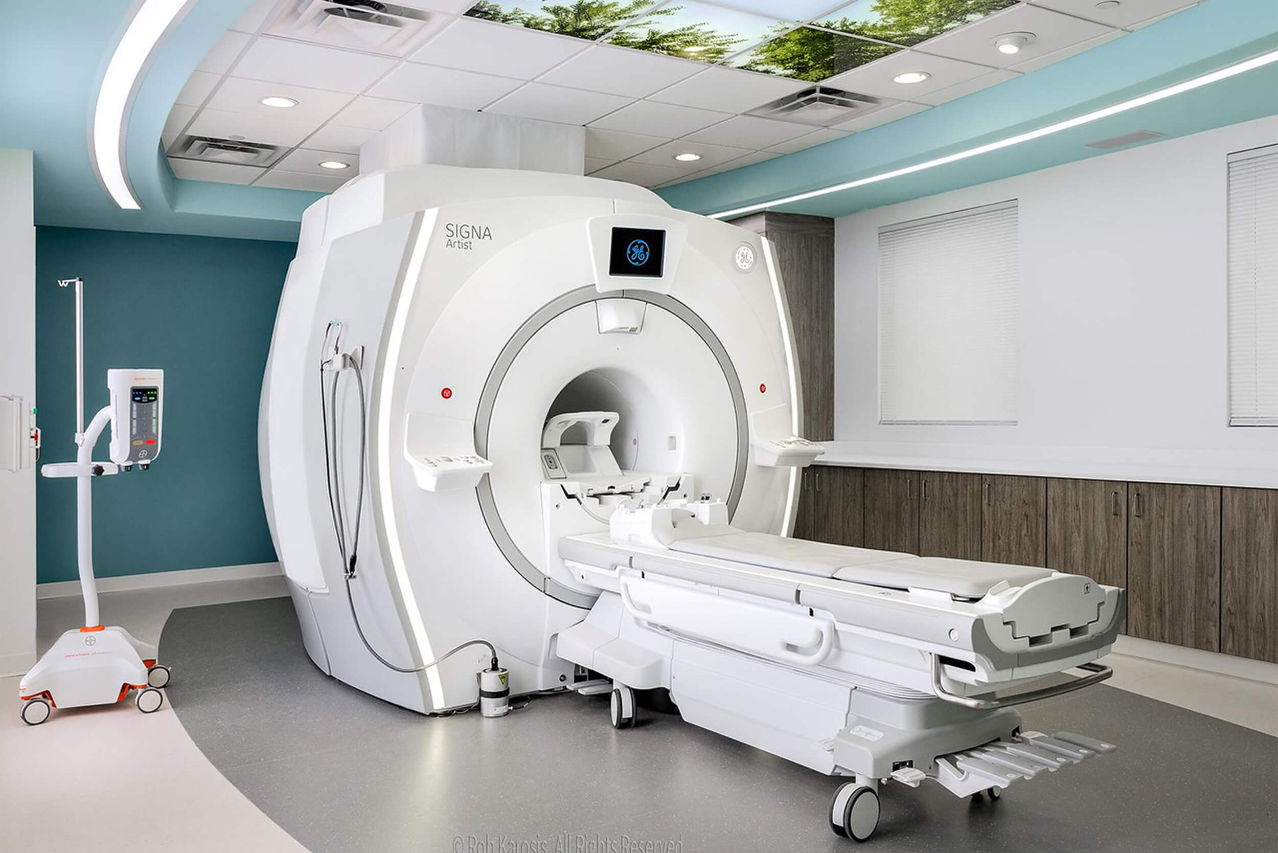 Interior view of an MRI imaging suite at a hospital. The room features medical equipment and a white MRI machine. The walls of the room are a light turquoise color.