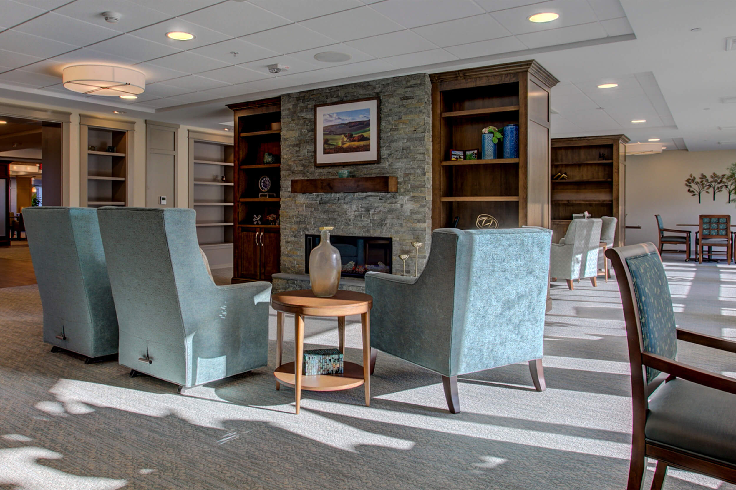 Interior view of living room area at a senior living facility. The room features neutral colored walls and grey wood flooring. Blue upholstered chairs and a small end table face a stone fireplace with bookcases on either side.