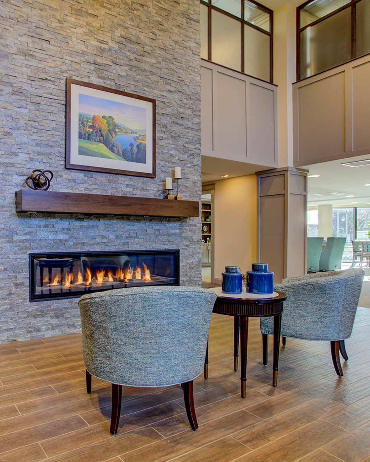 Interior view of the lobby of a senior living facility. A large stone fireplace column is seen with artwork, a minimalistic shelf, and modern horizontal fireplace is seen, along with two upholstered chairs and a small coffee table.