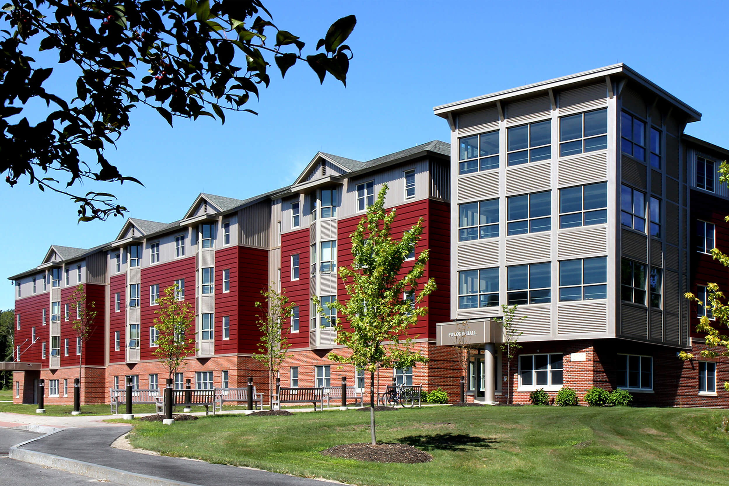 Exterior view of a college campus dorm building. The building facade features red and grey siding with brick along the bottom floor. A lawn with some planted trees are in front of the building.